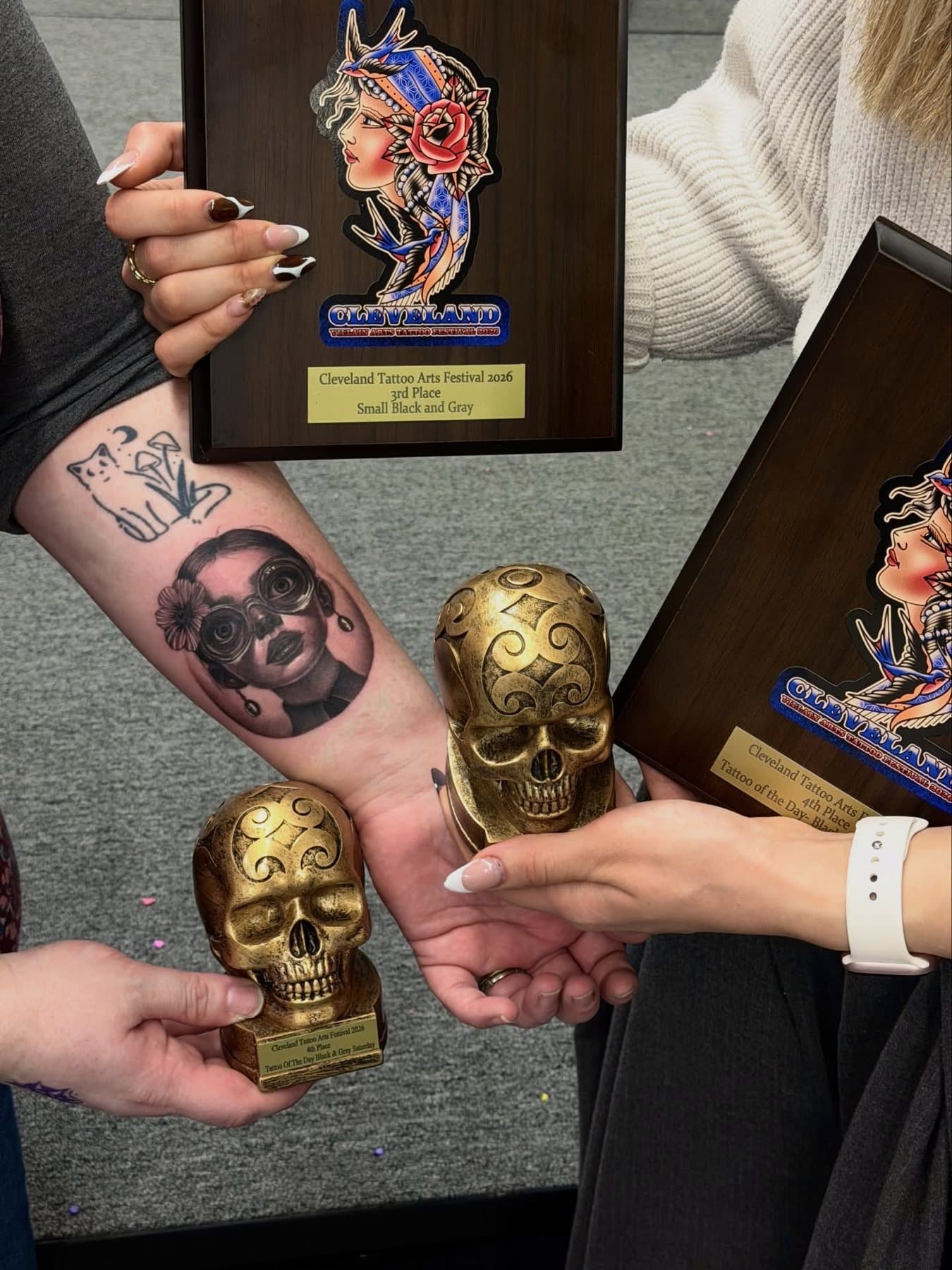 Cleveland Tattoo Arts Festival 2026 ödül plaketleri ve altın kuru kafa kupaları, siyah-gri mikro portre dövmesinin yanında