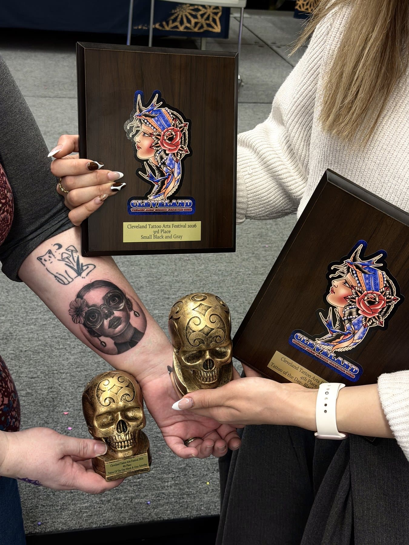 Ön kolda yuvarlak gözlüklü kız mikro portresi, iki Cleveland Tattoo Arts Festival 2026 plaketi ve kuru kafa kupalarıyla