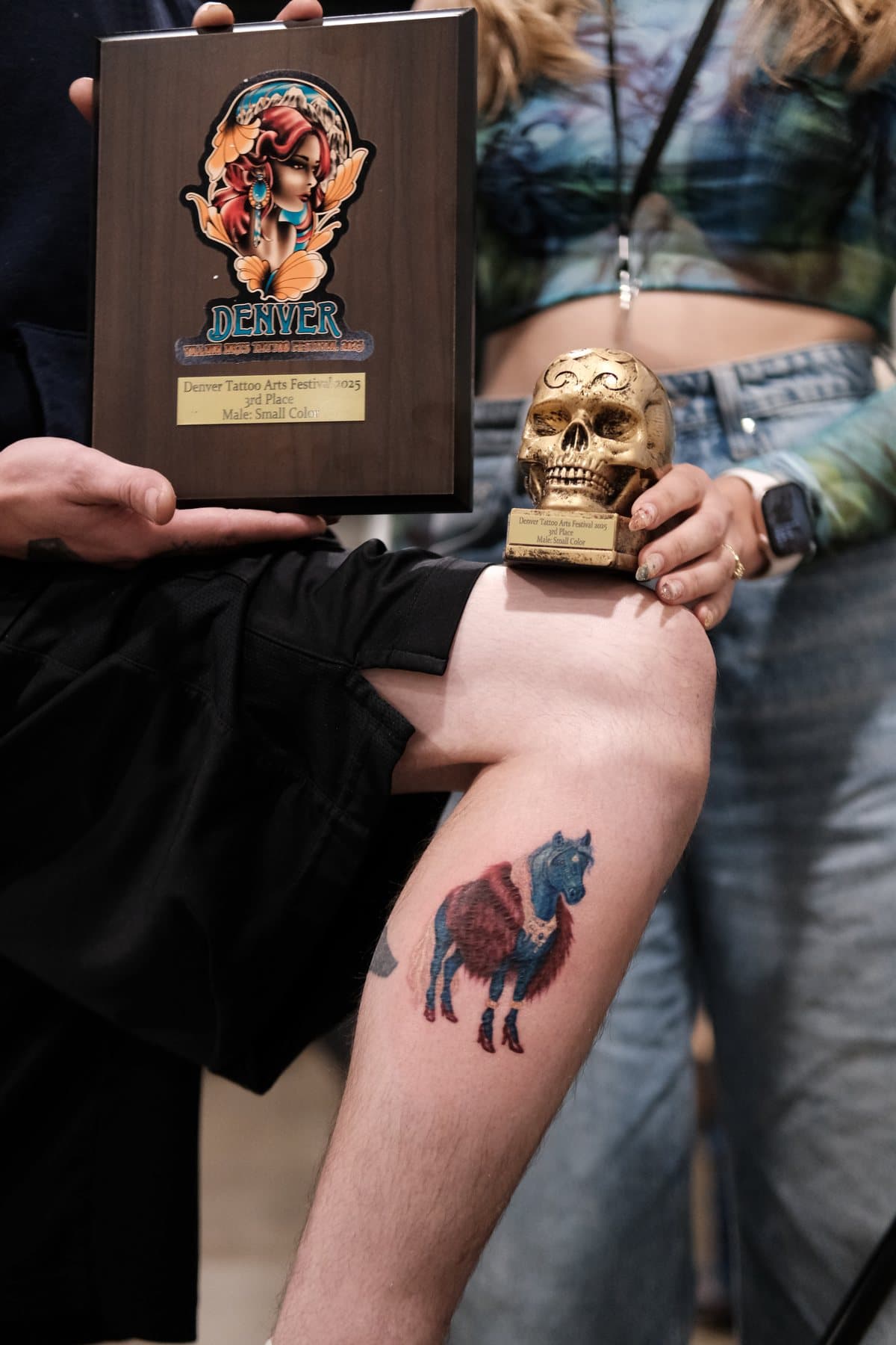 Baldırda kırmızı kürklü mavi at renkli dövmesi, Denver Tattoo Arts Festival 2025 plaketi ve kupasıyla