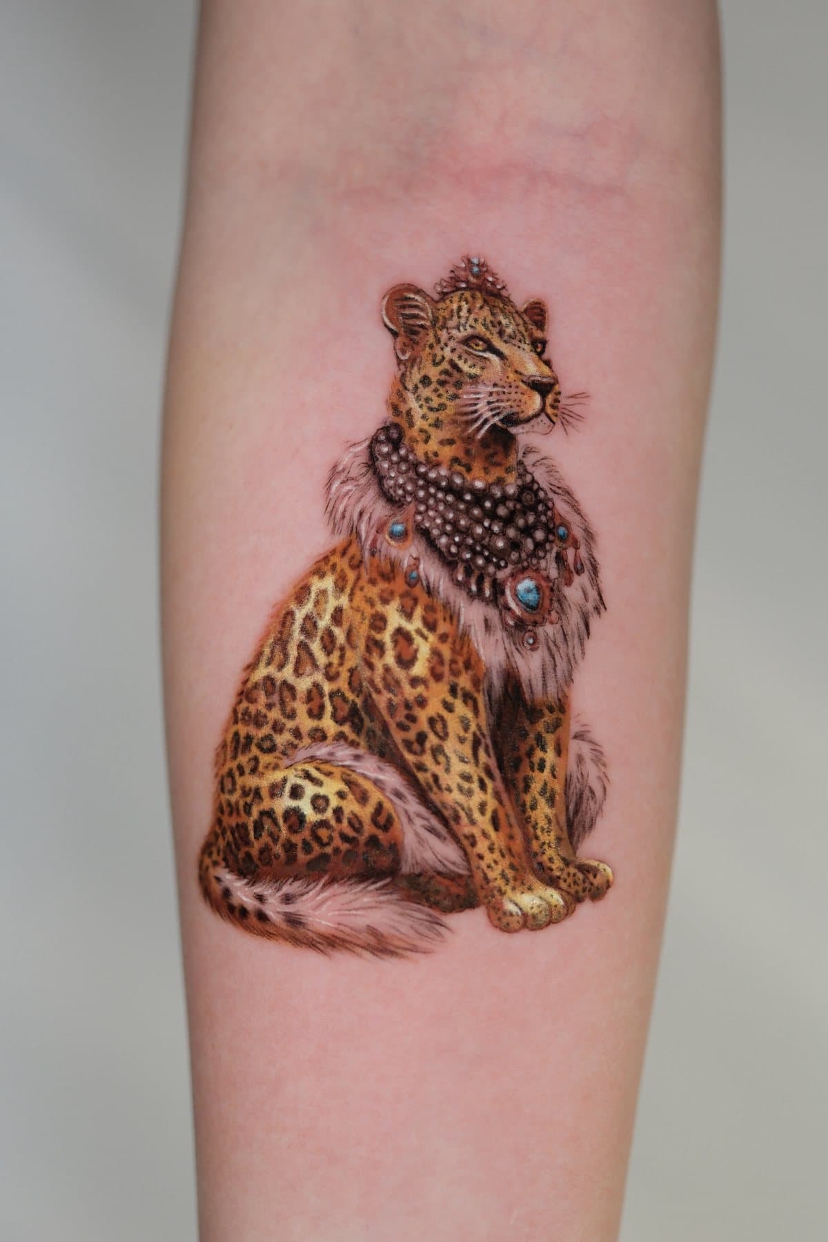 Color realism tattoo of a seated leopard wearing a tiara and layered pearl necklaces, on the forearm