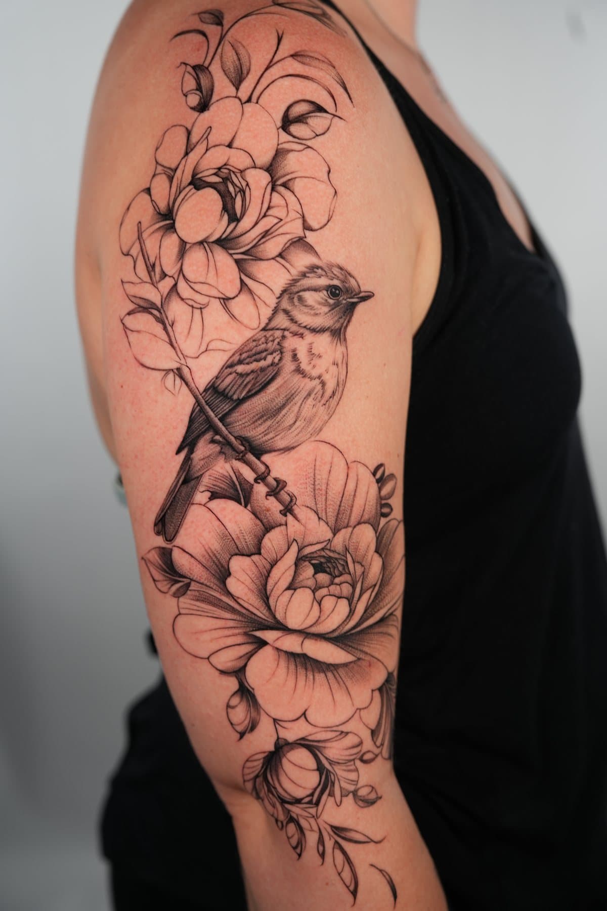 Black and grey fine line tattoo of a small songbird on a branch between large peonies, on the upper arm