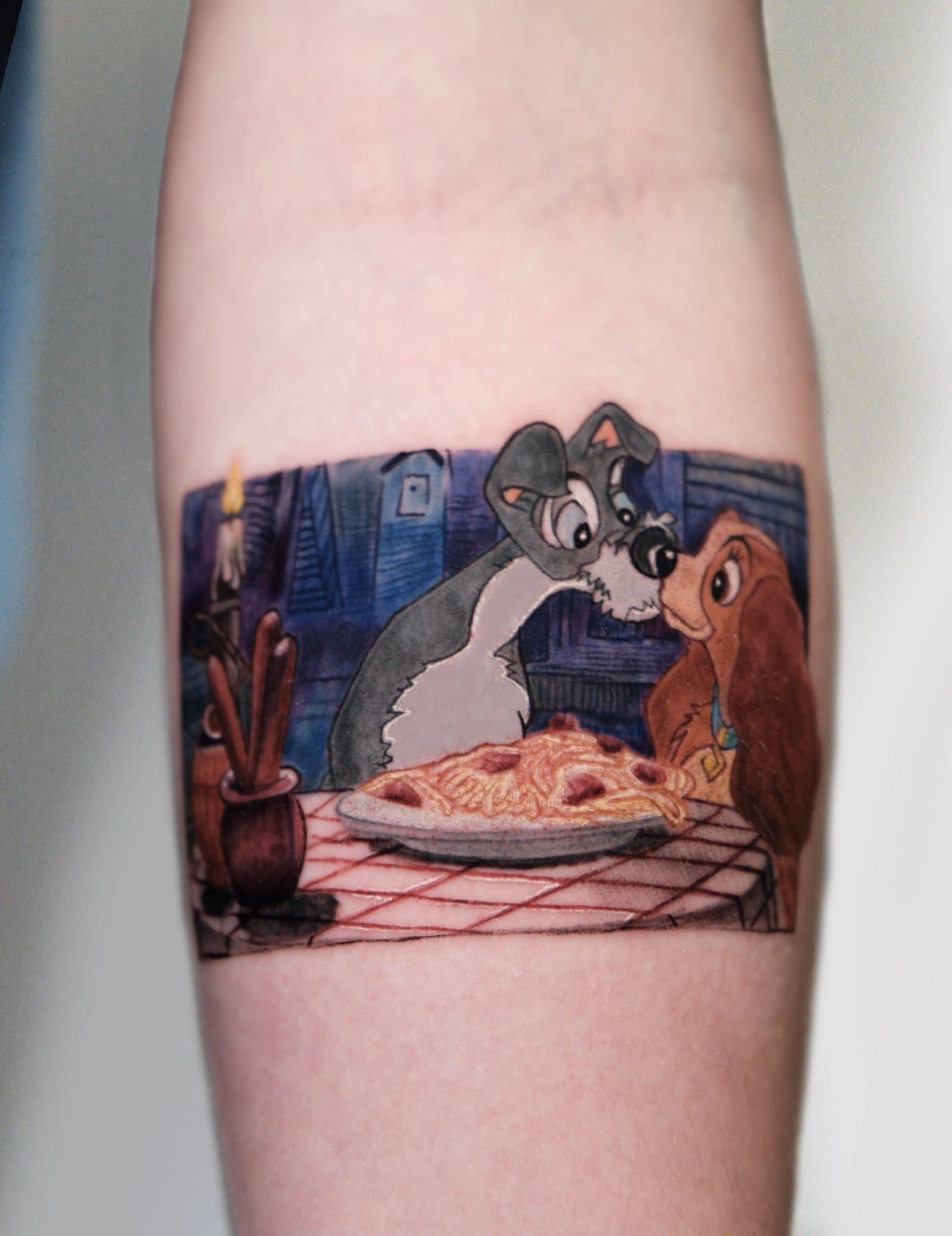 Color tattoo of the classic animated spaghetti dinner scene with two dogs sharing a plate, on the forearm