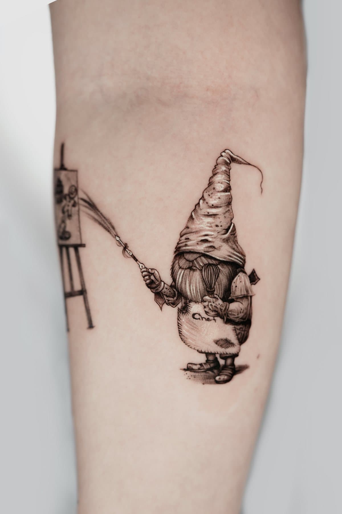 Black and grey micro tattoo of a gnome chef with a whisk, painting at an easel with a long brush
